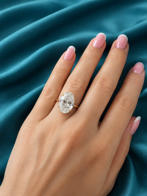 A close-up of a hand with pink manicured nails wearing a yellow gold ring with a large oval-cut clear gemstone, set against a draped teal satin fabric background.