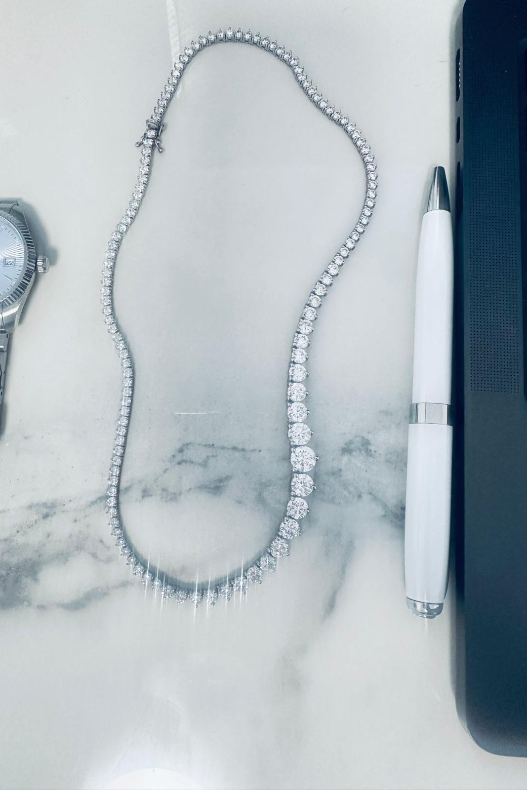 A white gold tennis necklace with a row of clear stones, displayed on a white marble in work setting